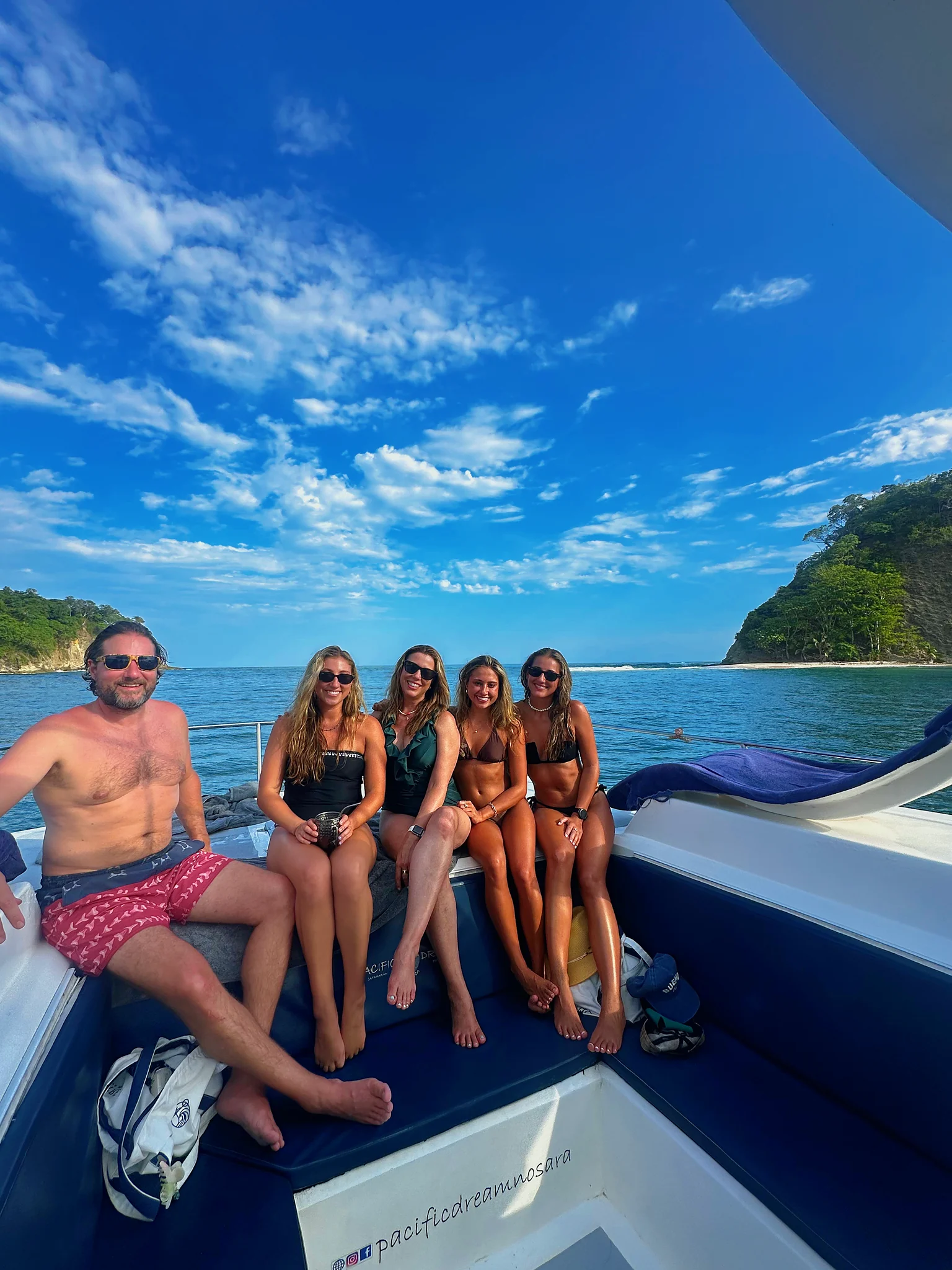 A group of friends smiles on the catamaran bow between two forested islands.