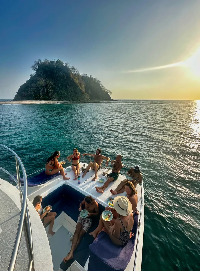 A group of friends relaxes on the catamaran bow as the boat approaches a green rocky island off Nosara.