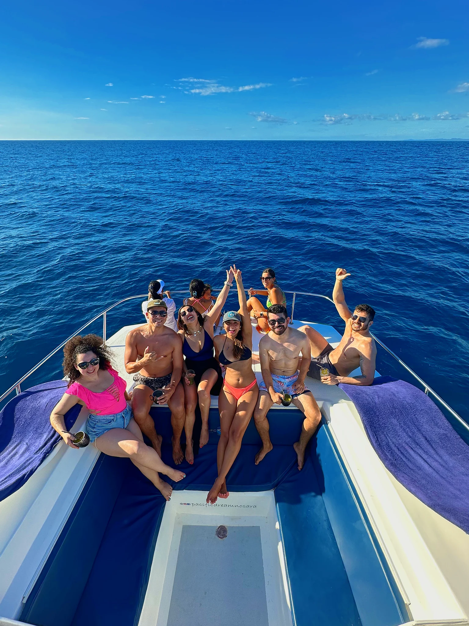 Friends wave and raise their drinks on the catamaran bow over deep blue water.