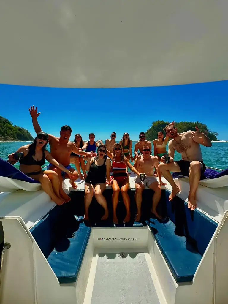 A large group cheers from the catamaran bow with a green island in the background.