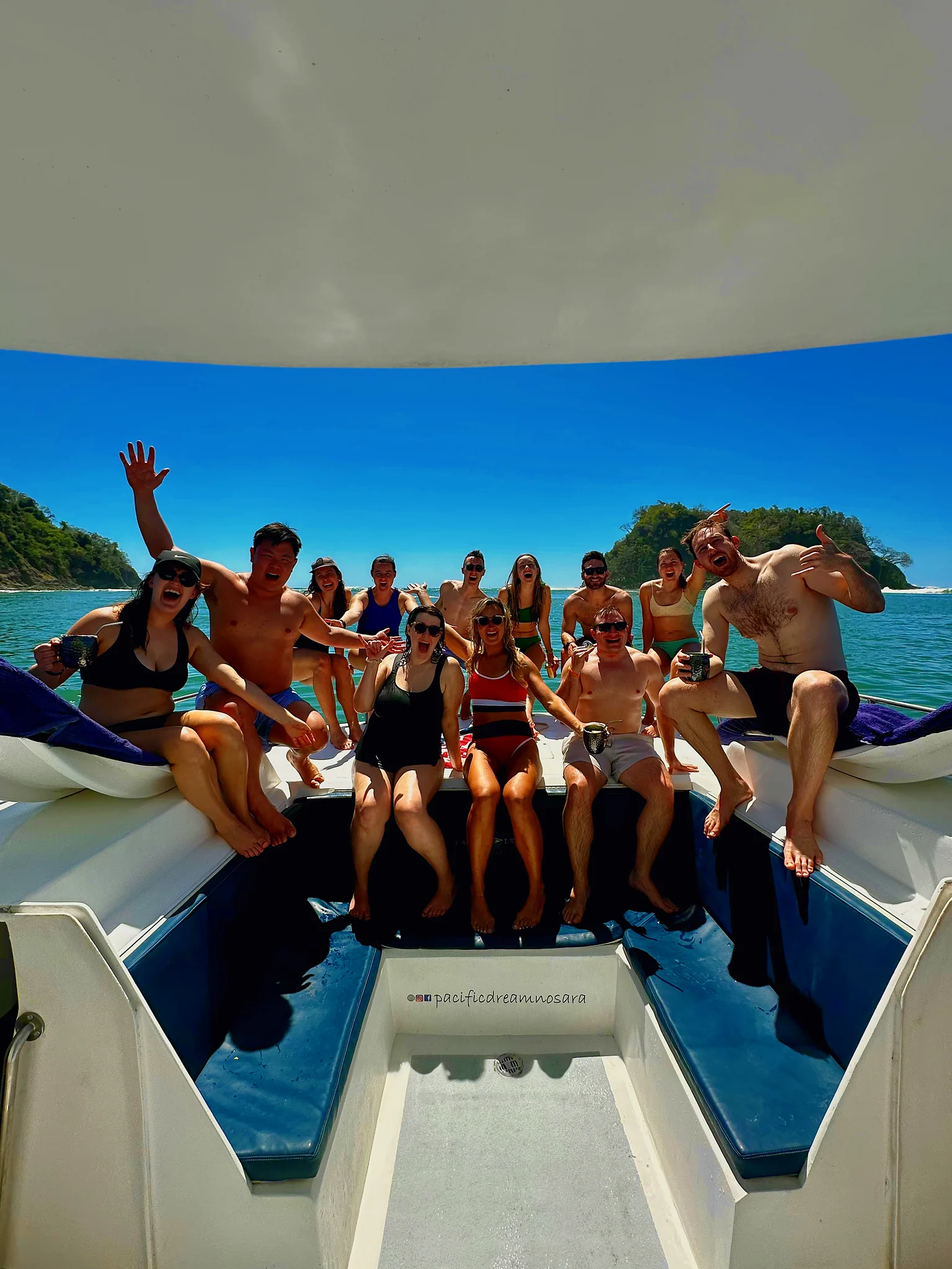 A large group cheers from the catamaran bow with a green island in the background.
