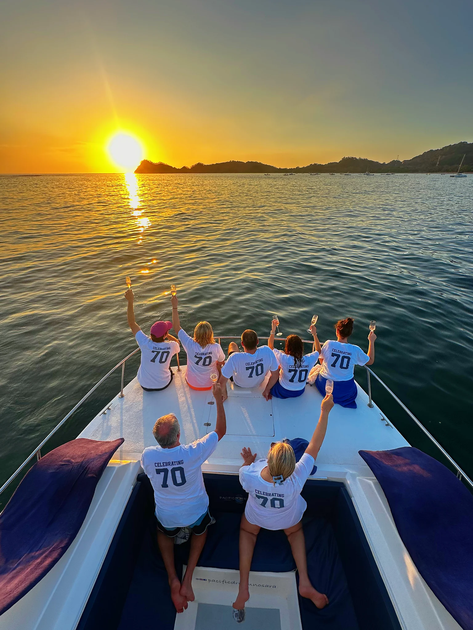 A group in matching birthday shirts cheers on the catamaran bow toward the setting sun.