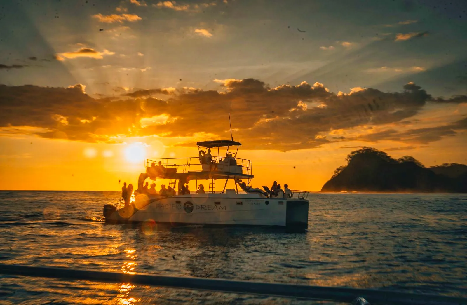 The Pacific Dream catamaran backlit by the setting sun beside a rocky island during the public sunset tour.