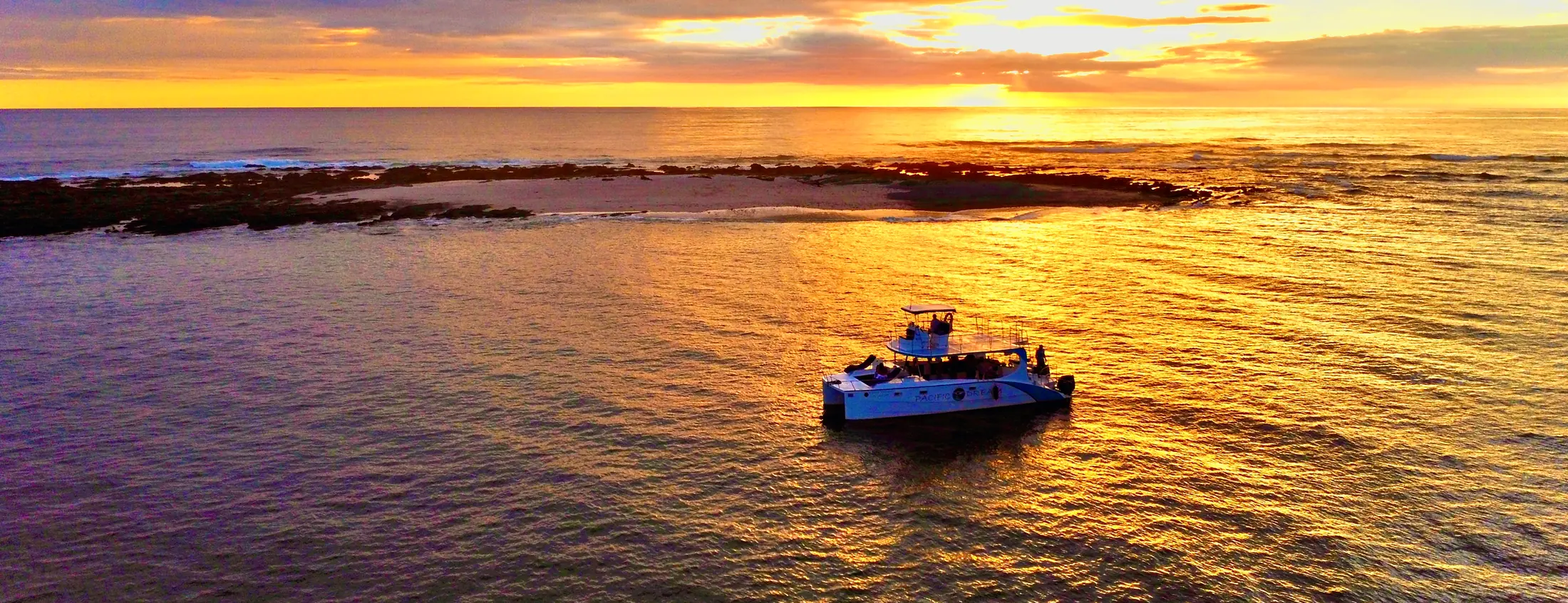Aerial view of the Pacific Dream catamaran anchored off a sandbar near Nosara as the sun sets over the Pacific.