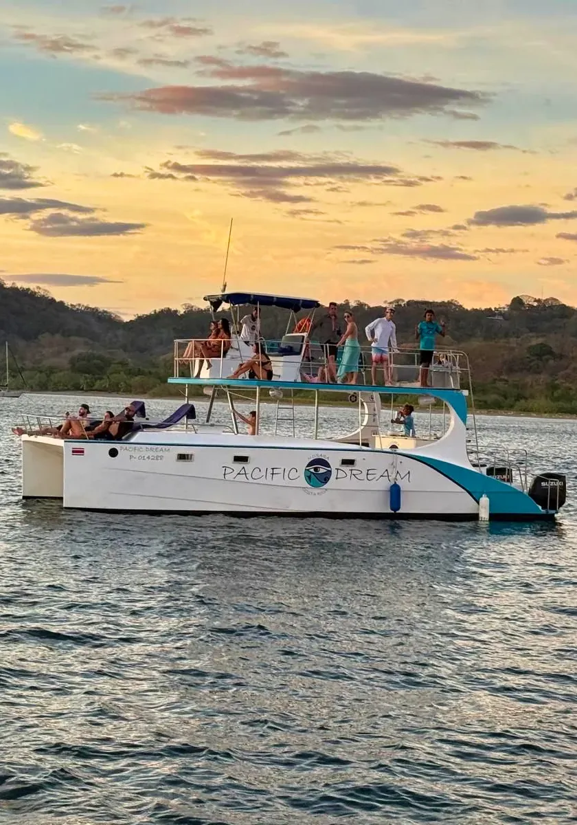 The Pacific Dream catamaran cruising at golden hour with guests on both decks and green hills behind.