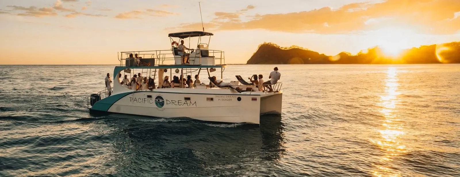 The Pacific Dream catamaran with guests on board, seen from the water as the sun sets behind the headland.