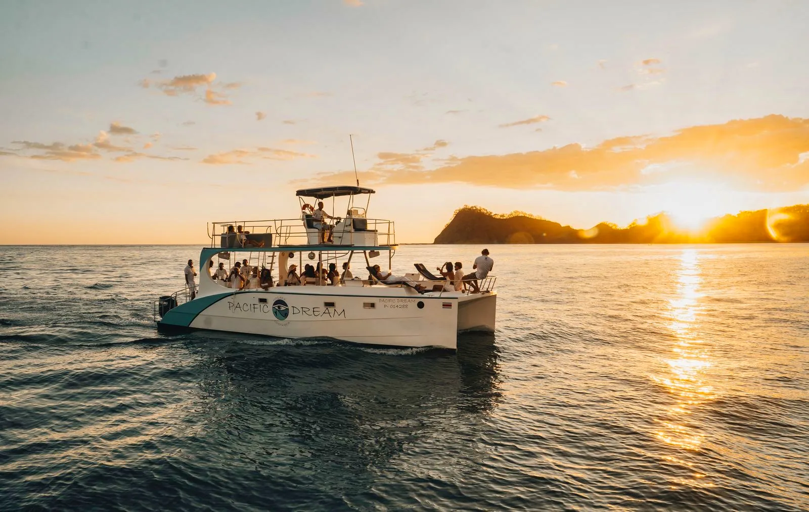 Pacific Dream catamaran seen from the water at sunset | Pacific Dream Nosara The Pacific Dream catamaran with guests on board, seen from the water as the sun sets behind the headland.