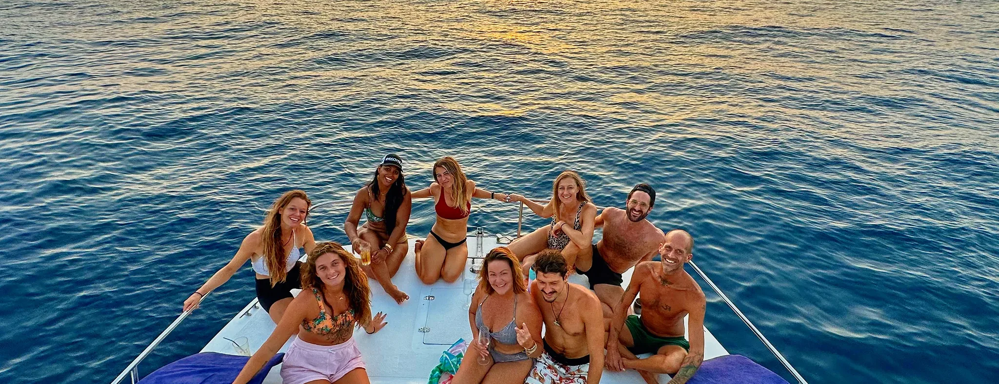A group of friends poses on the catamaran bow with the sun setting behind them